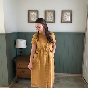 May include: A woman wearing a mustard yellow linen dress with a button-up front and pockets. She is standing in a room with green wainscoting and a wooden dresser.