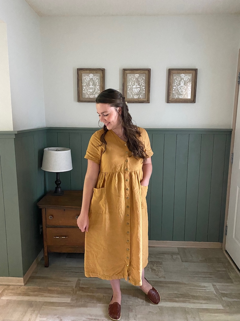 May include: A woman wearing a mustard yellow linen dress with a button-up front and pockets. She is standing in a room with green wainscoting and a wooden dresser.
