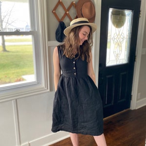 Pode incluir: Uma mulher vestindo um vestido de linho preto com bot&otilde;es na frente e bolsos. Ela est&aacute; usando um chap&eacute;u de palha e sorrindo.