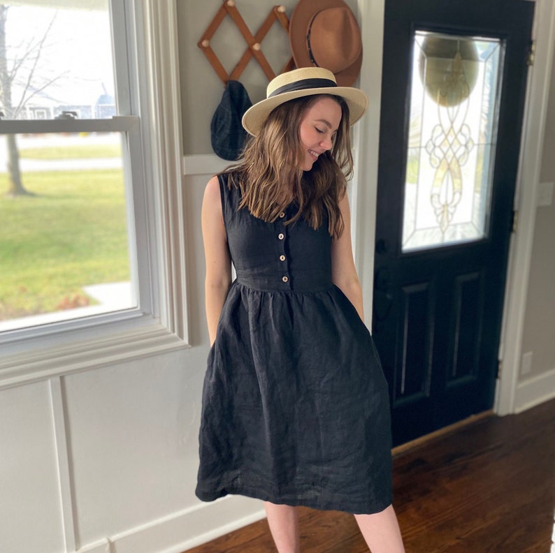 Pode incluir: Uma mulher vestindo um vestido de linho preto com bot&otilde;es na frente e bolsos. Ela est&aacute; usando um chap&eacute;u de palha e sorrindo.