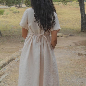 May include: A woman wearing a light beige linen dress with a tie at the waist. The dress has short sleeves and a flowy skirt.