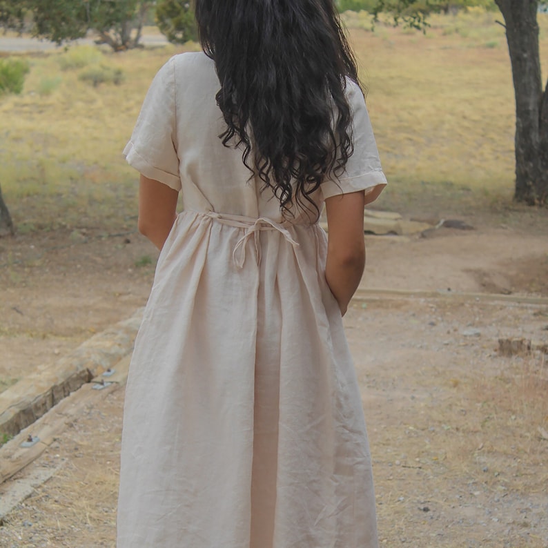 May include: A woman wearing a light beige linen dress with a tie at the waist. The dress has short sleeves and a flowy skirt.