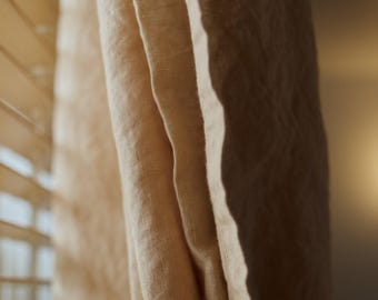 May include: A light beige curtain made of a textured fabric hangs in soft vertical folds. The material has a visible weave and is illuminated by natural light filtering through horizontal window blinds, creating shadows.