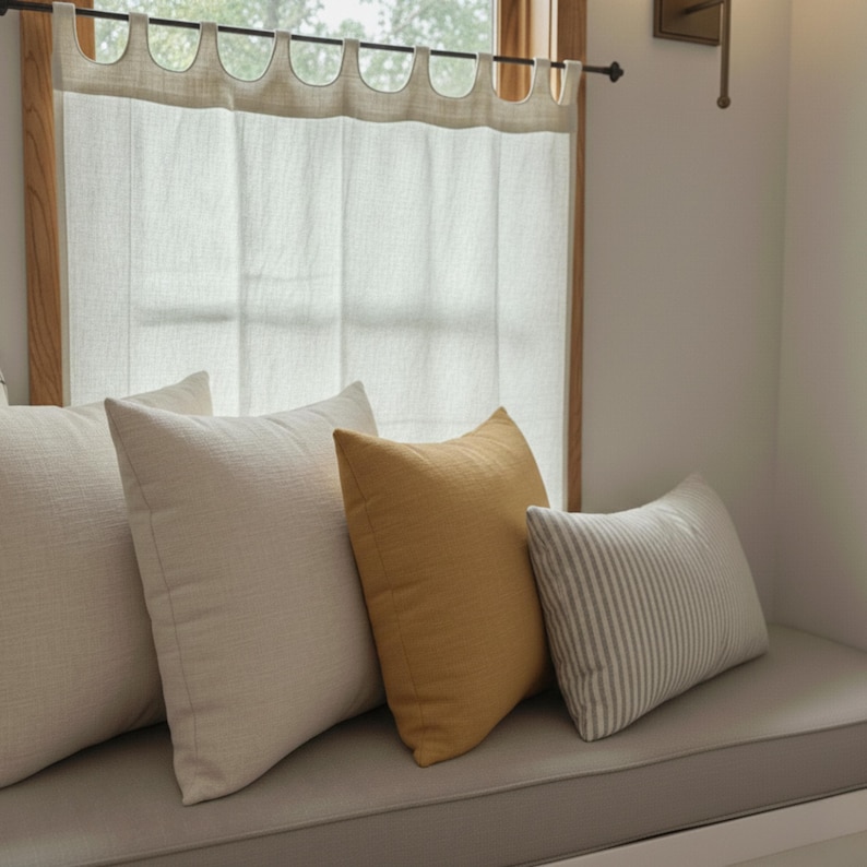 a cozy and inviting living space with a window, white curtains, and a comfortable couch adorned with three pillows in various colors and patterns.