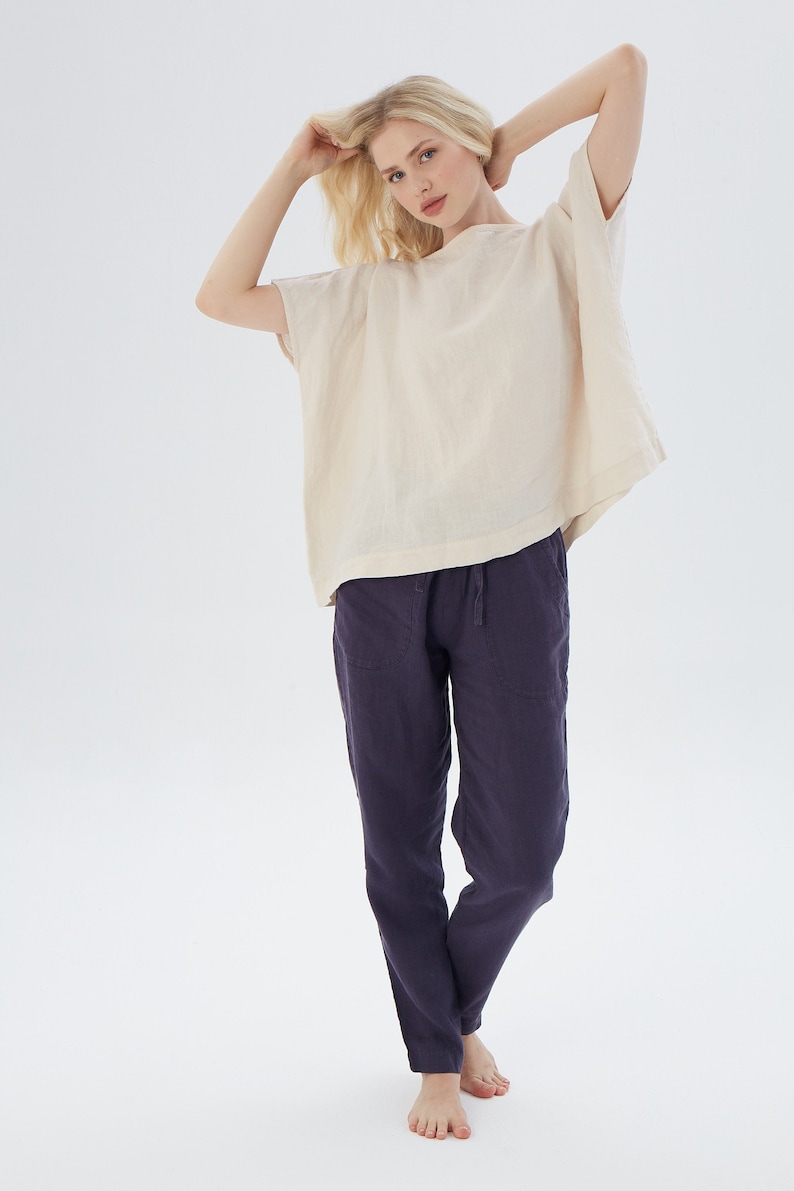 May include: A woman is wearing a loose-fitting, off-white linen top with short sleeves and a pair of dark purple linen pants. She is standing with her arms raised above her head, and her hair is blonde.