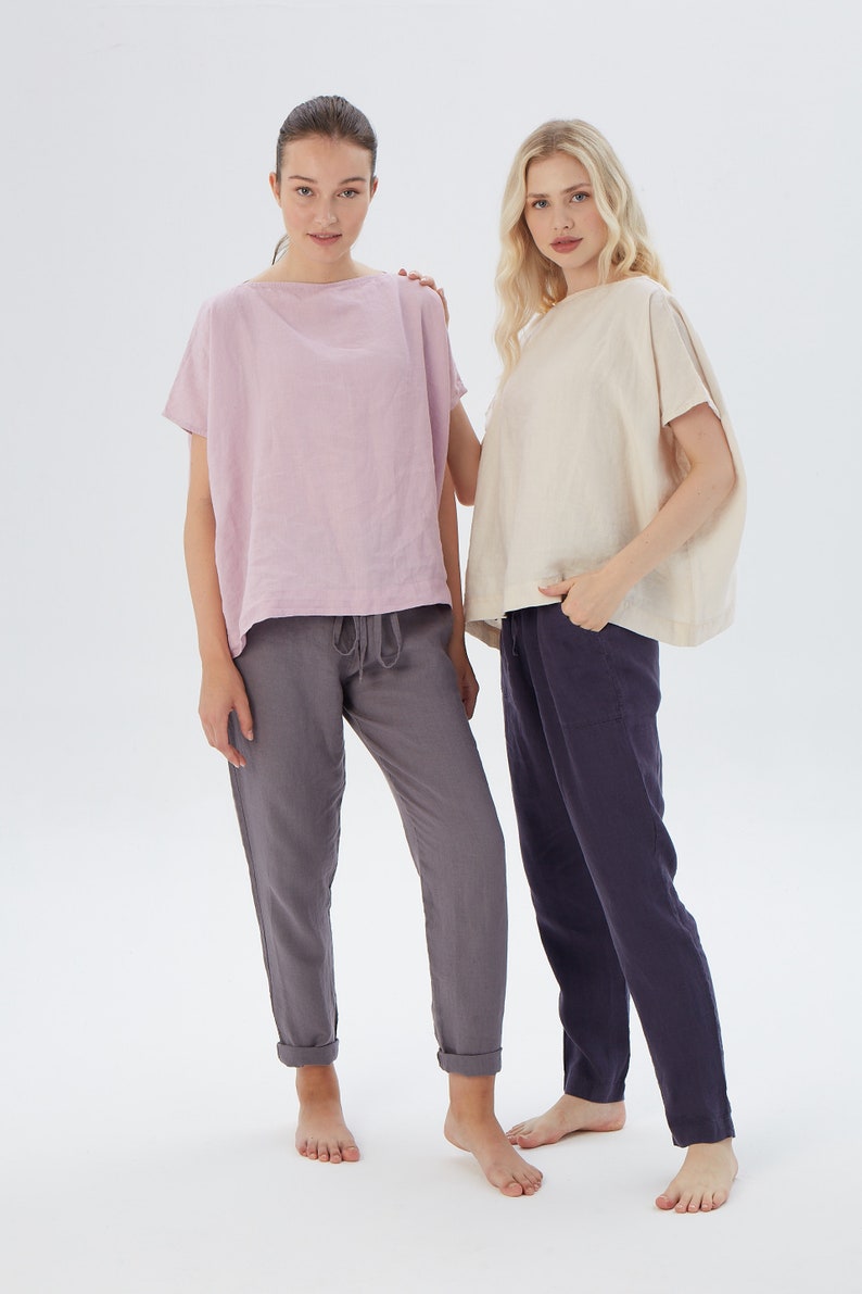 May include: Two women wearing loose-fitting linen tops and linen pants. The woman on the left is wearing a light pink top and gray pants. The woman on the right is wearing a cream-colored top and dark blue pants.
