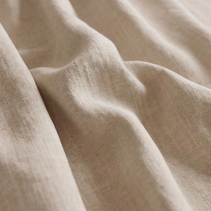 May include: A light beige fabric with a visible woven texture and a series of soft, deep folds. The material displays a natural, slightly irregular cross-hatch pattern throughout, creating a tactile appearance with varying light and shadow.