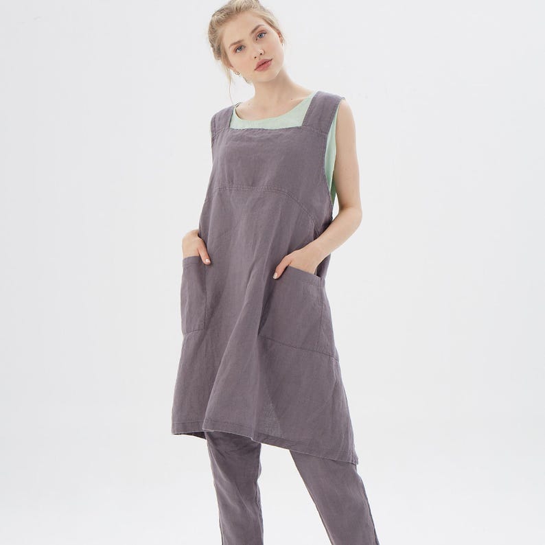 May include: A gray linen apron with two large pockets and a square neckline. The apron is sleeveless and has a loose fit.