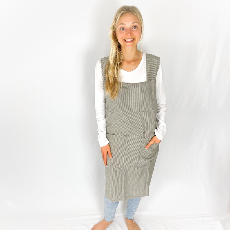 May include: A long, grey and white striped apron with a large pocket on the front. The apron is worn over a white long-sleeved shirt and blue jeans.