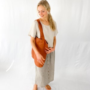 May include: A woman wearing a light beige linen top and a gray linen skirt with a row of buttons down the front. She is carrying a rust-colored tote bag.