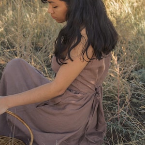 May include: A woman wearing a mauve-colored linen dress, seated in a field of tall, dry grass. The dress has a simple design with a tie-back detail. The overall aesthetic is natural and earthy, with a focus on the texture of the fabric and the outdoor setting.