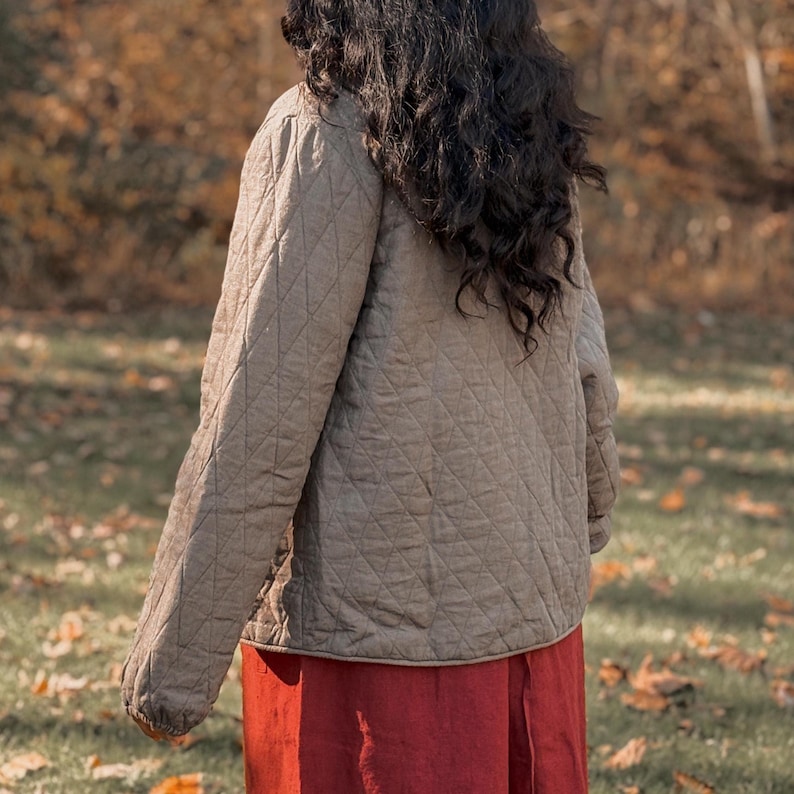Puede incluir: Chaqueta acolchada beige con un patr&oacute;n de diamantes, usada sobre una falda larga roja. Se ve el pelo oscuro de la persona. El fondo es una escena borrosa al aire libre con follaje oto&ntilde;al.
