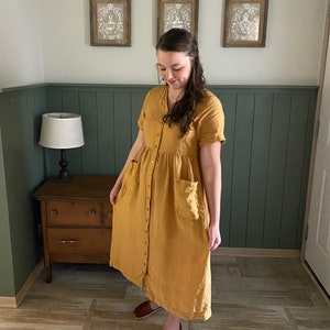 May include: A woman wearing a long, mustard yellow linen dress with a button-down front and pockets. She is standing on a wood-look tile floor in front of a green wainscot wall.