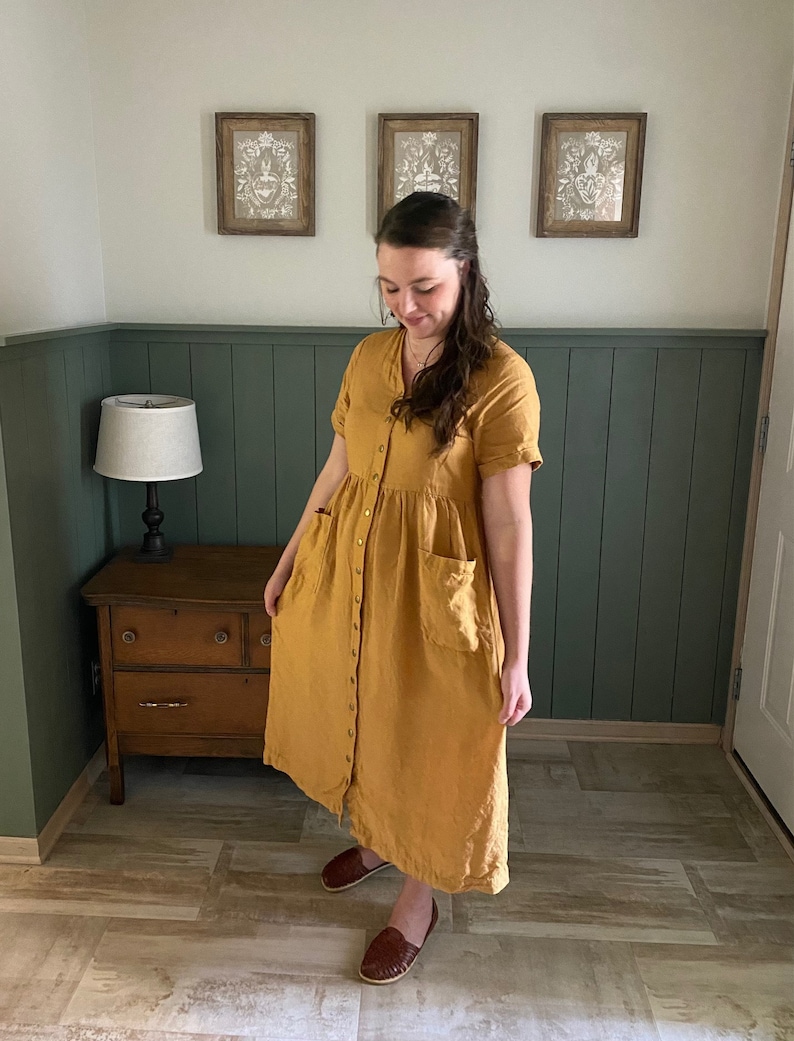 May include: A woman wearing a long, mustard yellow linen dress with a button-down front and pockets. She is standing on a wood-look tile floor in front of a green wainscot wall.