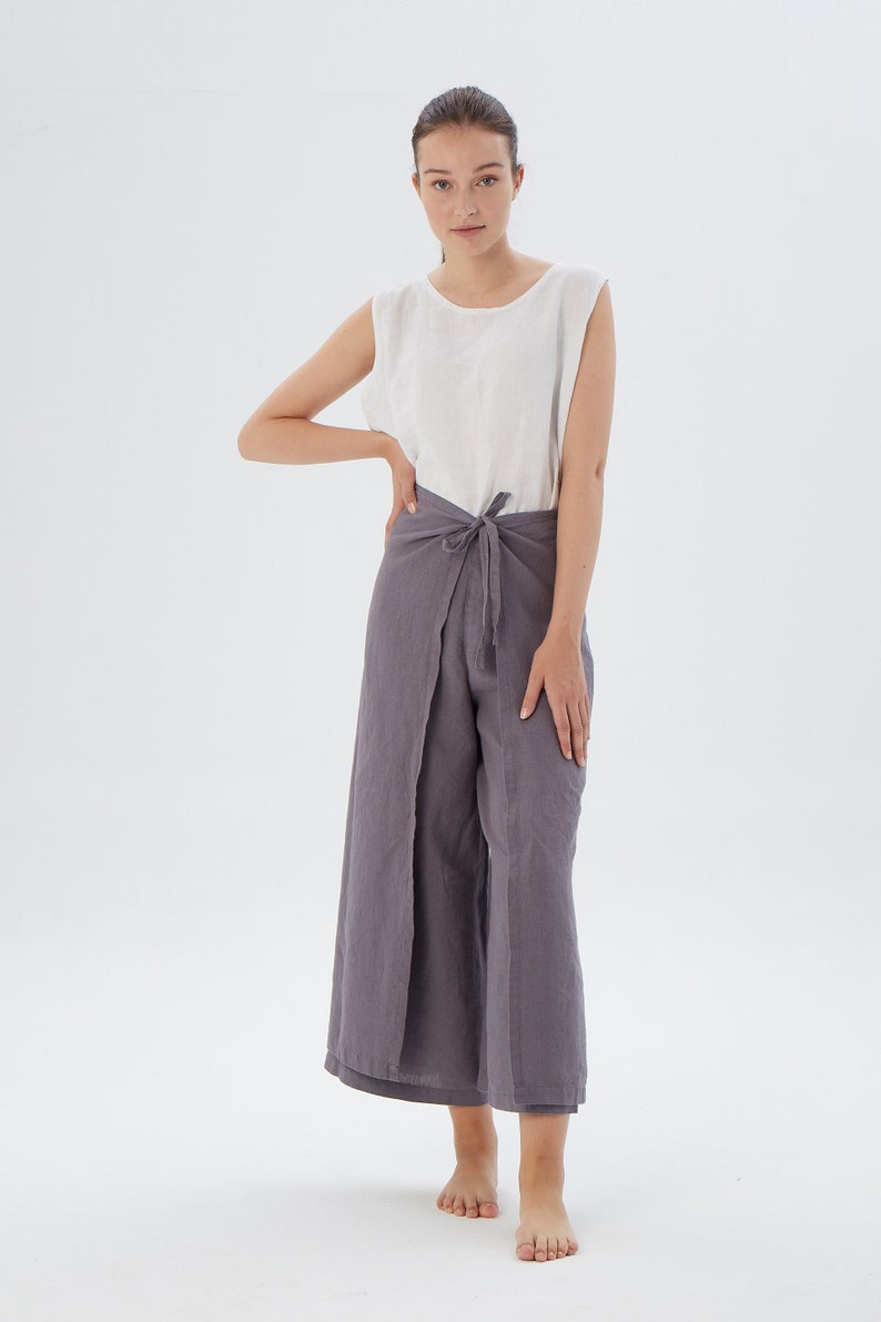 Puede incluir: Una mujer con una camiseta sin mangas blanca y pantalones envolventes grises. Los pantalones se atan a la cintura y tienen un dise&ntilde;o de pierna ancha. El atuendo est&aacute; hecho de una tela suave y fluida, perfecto para el clima c&aacute;lido. El fondo es blanco liso.