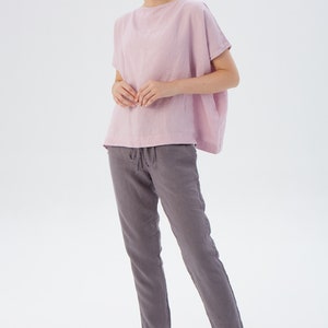 May include: A woman is wearing a light pink linen top with short sleeves and a pair of gray linen pants. The pants have a drawstring waist.