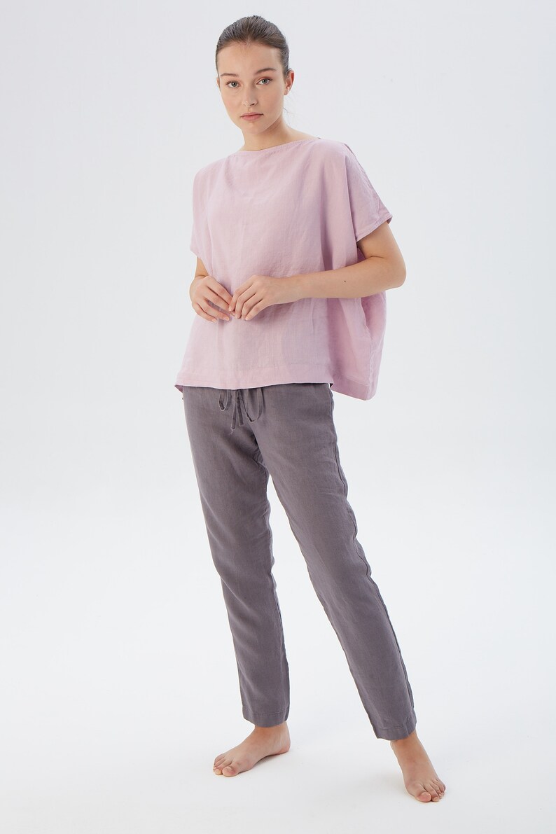 May include: A woman is wearing a light pink linen top with short sleeves and a pair of gray linen pants. The pants have a drawstring waist.