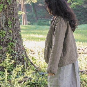 May include: A person wearing a quilted olive green jacket and a light gray striped skirt stands in a sunlit forest. The jacket has a button closure and pockets. The skirt is a midi length.