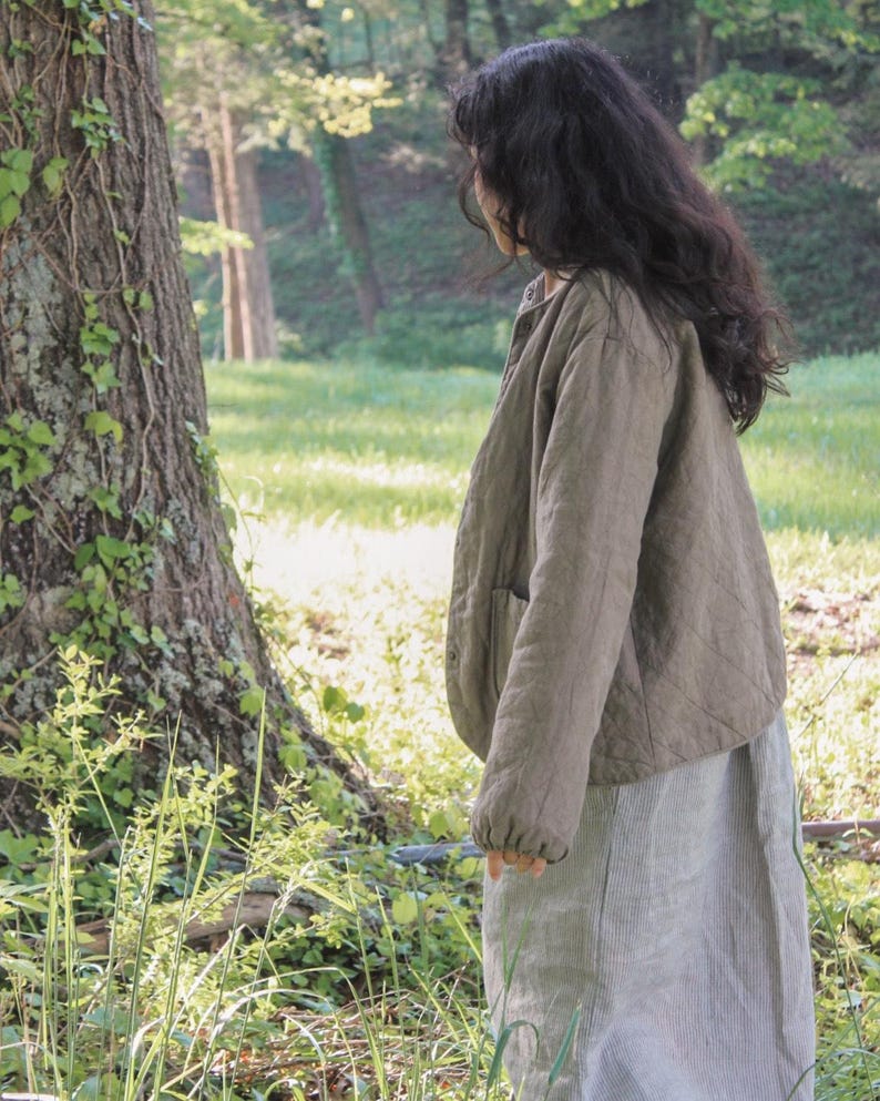 May include: A person wearing a quilted olive green jacket and a light gray striped skirt stands in a sunlit forest. The jacket has a button closure and pockets. The skirt is a midi length.