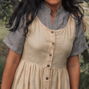 May include: A beige linen dress with a button-up front and a gray striped shirt underneath. The dress has a gathered skirt and a relaxed fit.