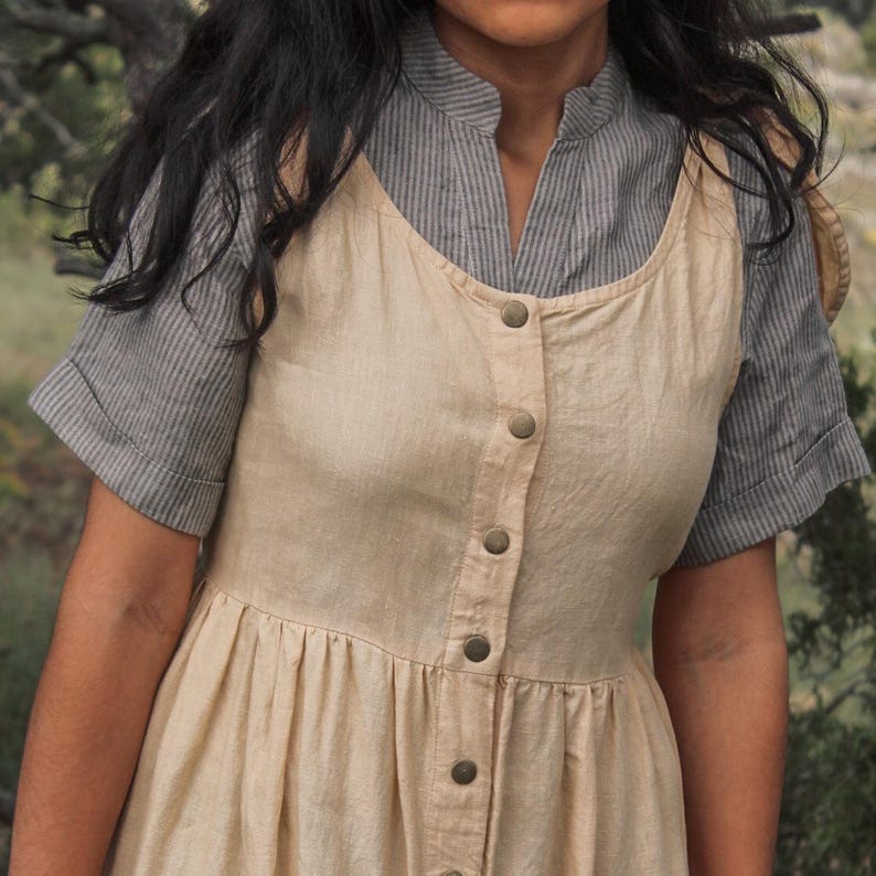 May include: A beige linen dress with a button-up front and a gray striped shirt underneath. The dress has a gathered skirt and a relaxed fit.