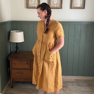 May include: A woman wearing a yellow linen dress with a button-down front and pockets. She is wearing brown leather sandals.