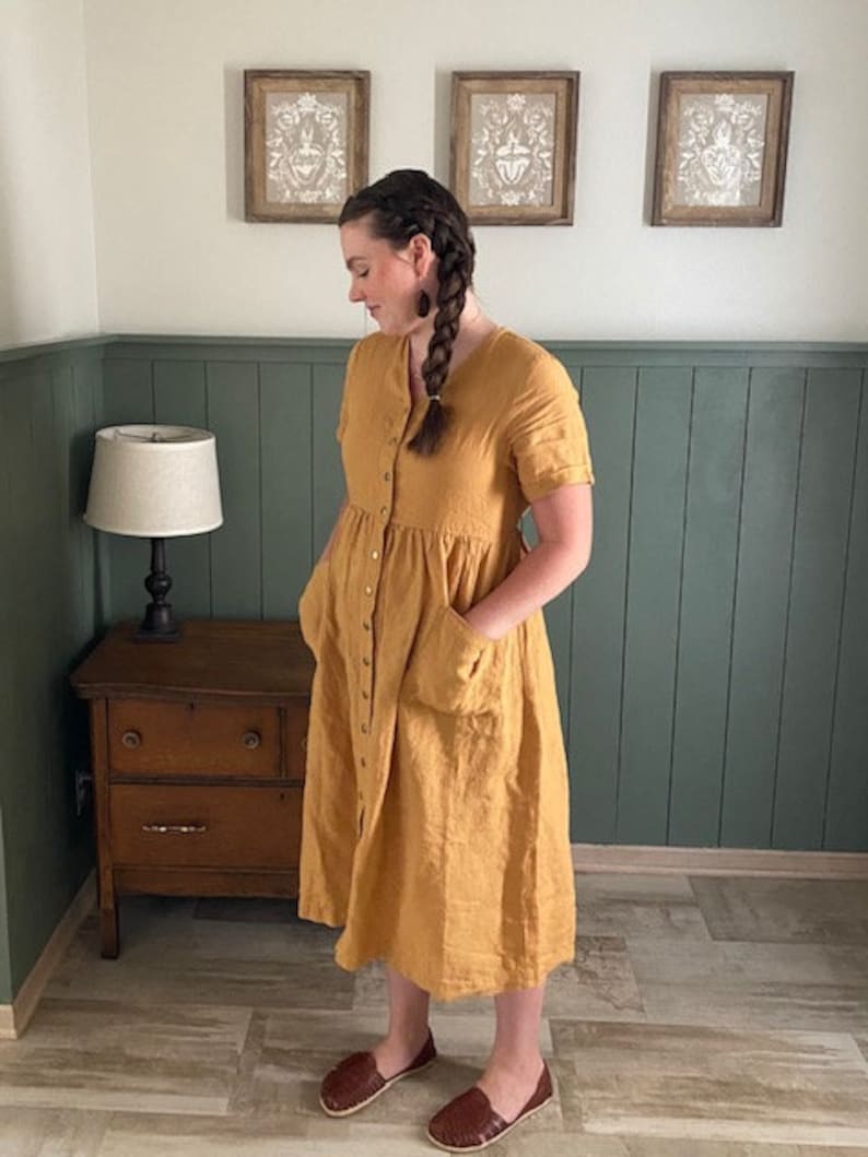 May include: A woman wearing a yellow linen dress with a button-down front and pockets. She is wearing brown leather sandals.