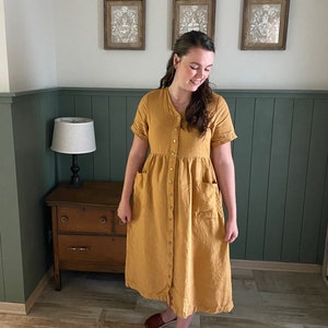 May include: A woman wearing a mustard yellow linen dress with a button-up front and pockets. She is standing in a room with green wainscoting and a wooden dresser.