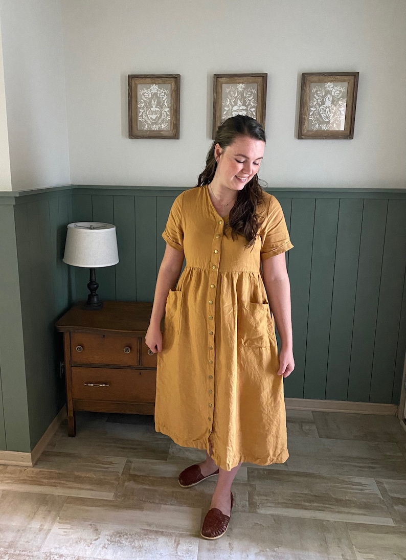 May include: A woman wearing a mustard yellow linen dress with a button-up front and pockets. She is standing in a room with green wainscoting and a wooden dresser.