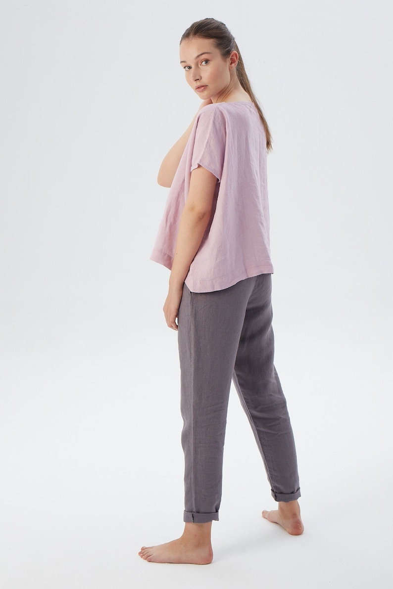May include: A woman is wearing a loose-fitting, light pink linen top with short sleeves and a pair of dark gray linen pants. She is standing with her back to the camera and looking over her shoulder.