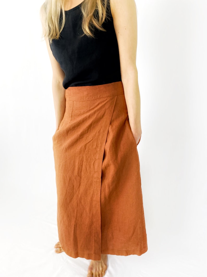 May include: A rust-colored linen wrap skirt with a long, flowing silhouette. The skirt has a simple design with a single wrap and a tie at the waist.