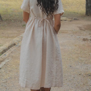 May include: A woman wearing a long, light beige linen dress with a tie at the waist. She is wearing brown leather boots and standing on a wooden platform.