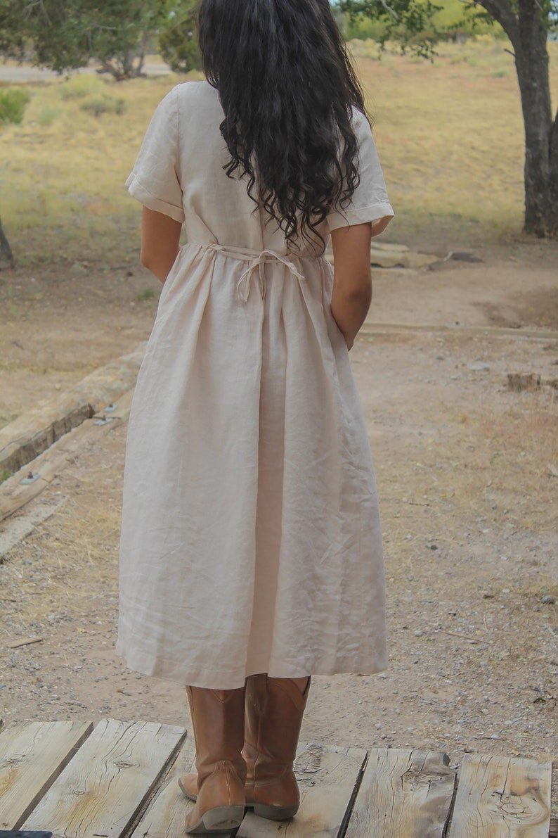 May include: A woman wearing a long, light beige linen dress with a tie at the waist. She is wearing brown leather boots and standing on a wooden platform.