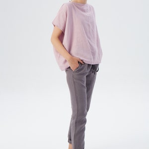 May include: A woman wearing a light pink linen top with short sleeves and a pair of gray linen pants. She is standing with her hands in her pockets and looking at the camera.