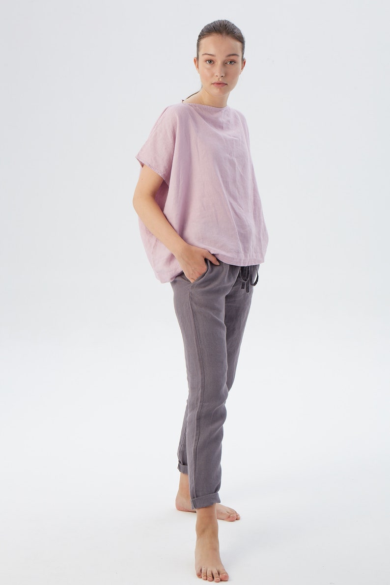 May include: A woman wearing a light pink linen top with short sleeves and a pair of gray linen pants. She is standing with her hands in her pockets and looking at the camera.