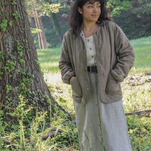 May include: A woman wearing a light olive green quilted jacket with a button-down dress. The dress is a light gray and white striped pattern. The woman is standing in a grassy area with a large tree.