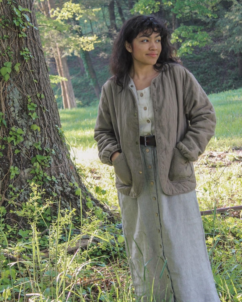 May include: A woman wearing a light olive green quilted jacket with a button-down dress. The dress is a light gray and white striped pattern. The woman is standing in a grassy area with a large tree.