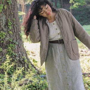 May include: A woman wearing a light brown quilted jacket with a round collar and two front pockets. She is also wearing a white button-down shirt, a striped button-down skirt, and a brown belt. The outfit is set in a natural outdoor setting.