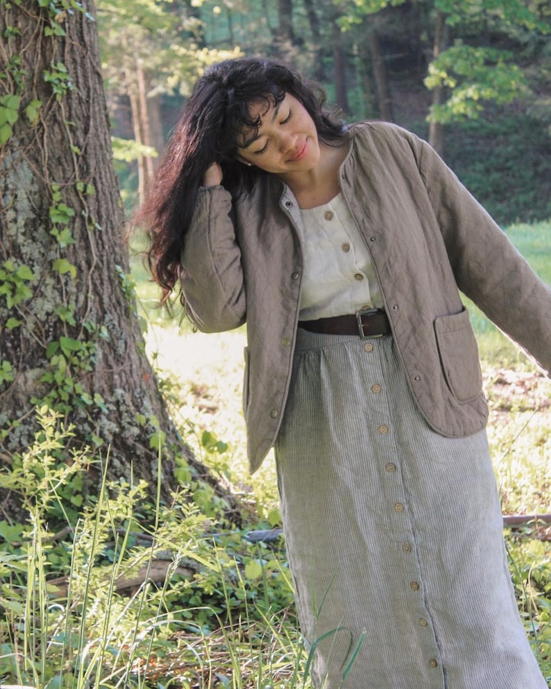 May include: A woman wearing a light brown quilted jacket with a round collar and two front pockets. She is also wearing a white button-down shirt, a striped button-down skirt, and a brown belt. The outfit is set in a natural outdoor setting.