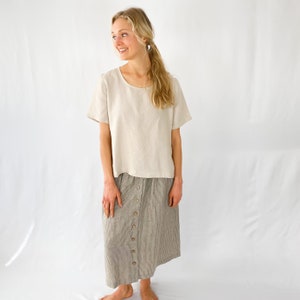 May include: A woman wearing a light beige linen top with short sleeves and a gray and white striped linen skirt with a button closure.
