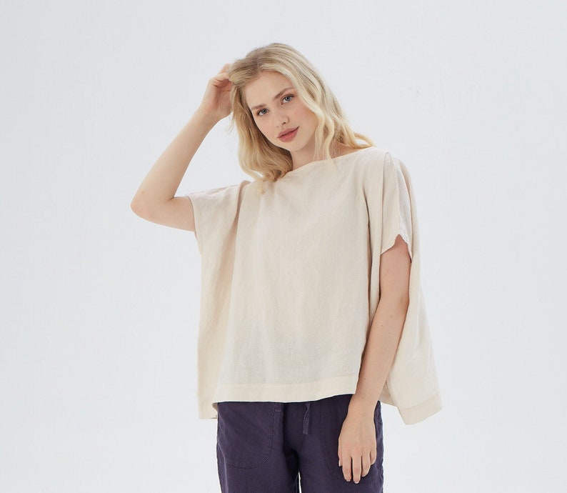 May include: A woman wearing a loose-fitting, off-white linen top with short sleeves and a boat neck. She is wearing dark purple pants.