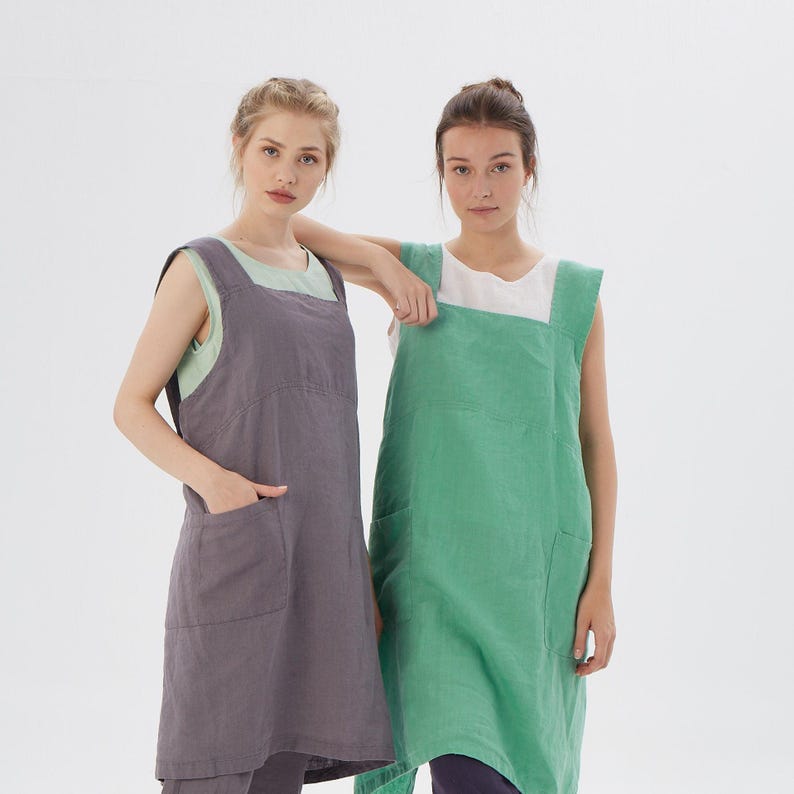 May include: Two women wearing linen aprons. The apron on the left is a dark gray color and the apron on the right is a light green color. Both aprons have a single pocket on each side.