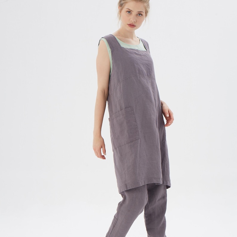 May include: A long, sleeveless, gray linen apron with a large pocket on the front. The apron is worn over a light green shirt and gray linen pants.
