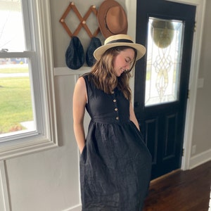 Pode incluir: Uma mulher vestindo um vestido de linho preto com bot&otilde;es na frente e bolsos. Ela est&aacute; usando um chap&eacute;u de palha com uma faixa preta.