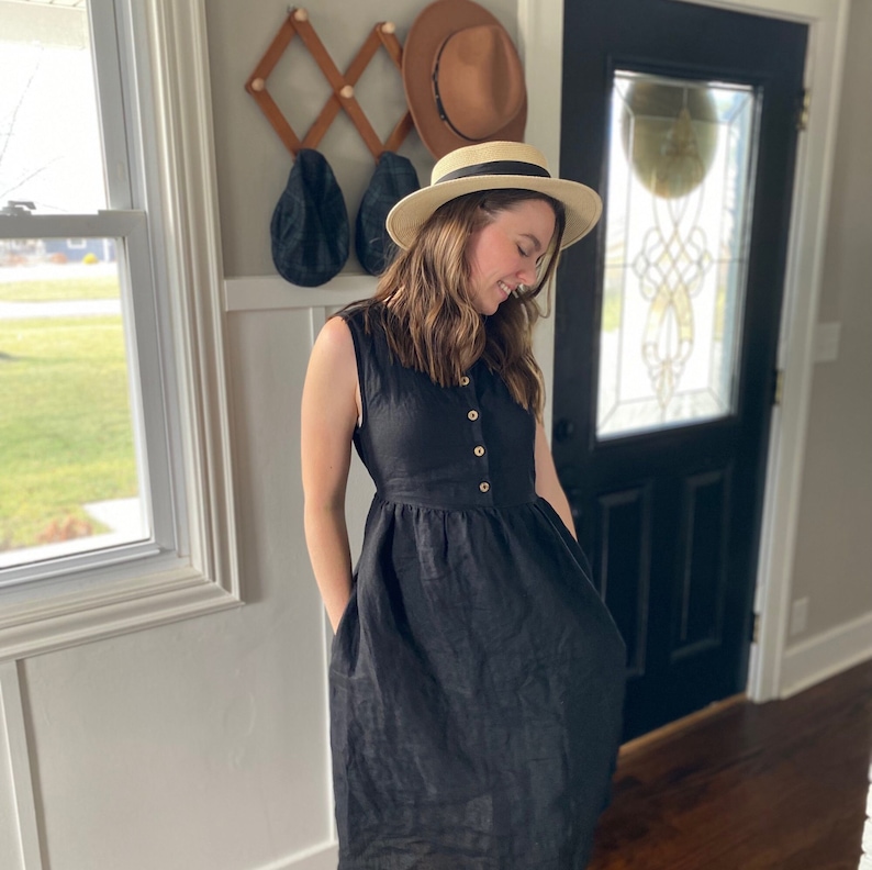 Pode incluir: Uma mulher vestindo um vestido de linho preto com bot&otilde;es na frente e bolsos. Ela est&aacute; usando um chap&eacute;u de palha com uma faixa preta.