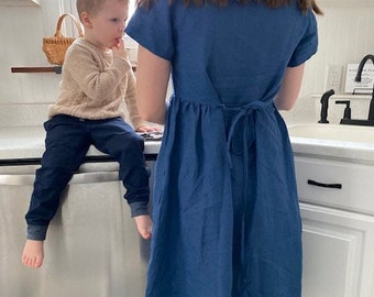 Puede incluir: Una mujer lleva un vestido de lino azul con un lazo en la espalda. Un niño pequeño está de pie junto a ella, con un suéter beige y pantalones azules.