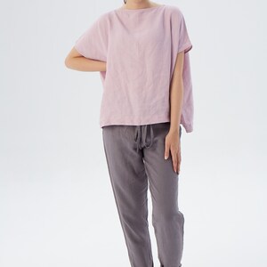 May include: A woman is wearing a loose-fitting, pale pink linen top with short sleeves and a pair of dark gray linen pants. The pants have a drawstring waist and are cuffed at the ankles.