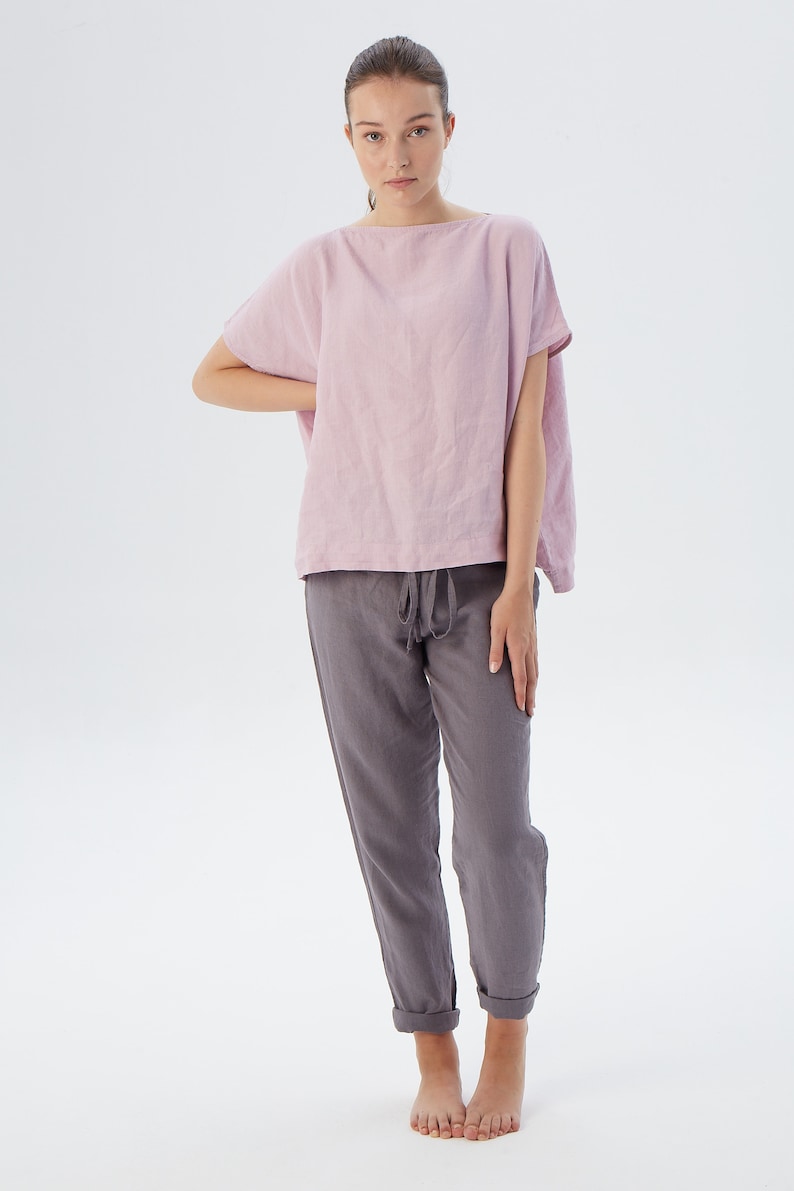 May include: A woman is wearing a loose-fitting, pale pink linen top with short sleeves and a pair of dark gray linen pants. The pants have a drawstring waist and are cuffed at the ankles.