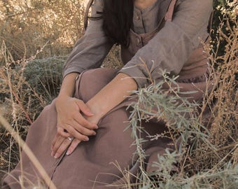 May include: A woman wearing a brown linen dress with a ruffled hem sits in a field of tall grass. She is wearing a simple silver ring on her finger.
