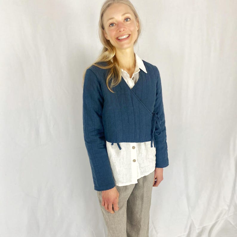 May include: A dark blue wrap jacket with long sleeves and a tie closure. The jacket is made of a soft, quilted fabric and is worn over a white button-down shirt and brown pants.